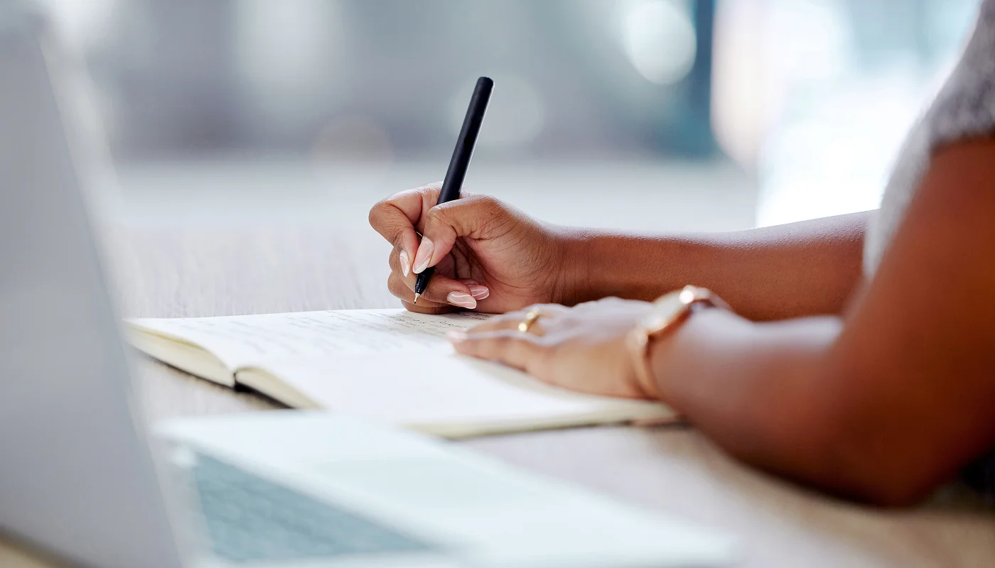 person making notes in a notebook on their desk with laptop in the foreground